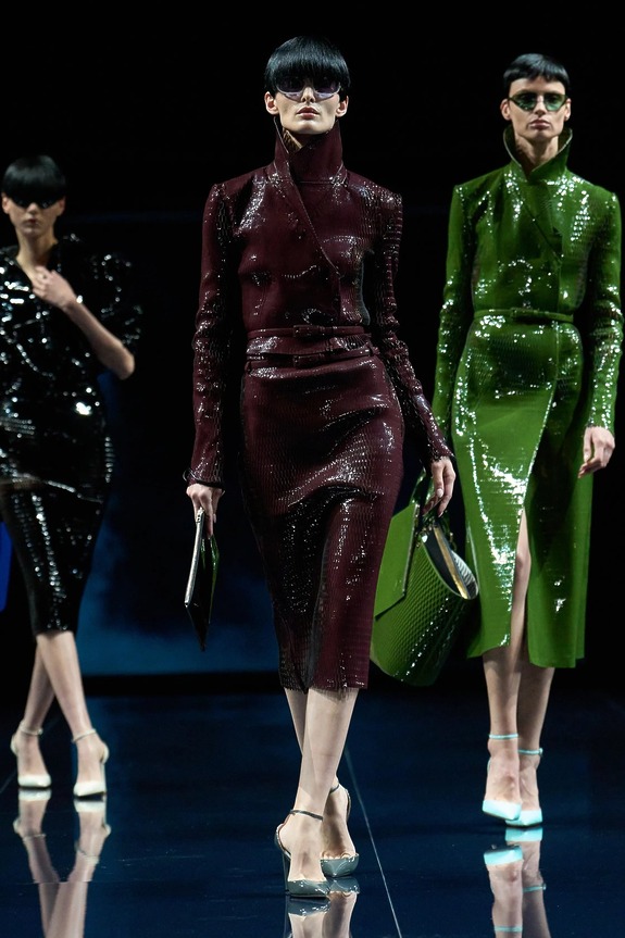 Models walking the runway in stylish, shiny outfits featuring dark red, black, and green designs with futuristic eyewear.