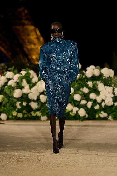Model wearing a blue, shiny outfit and sunglasses, walking on a runway with white flowers in the background.