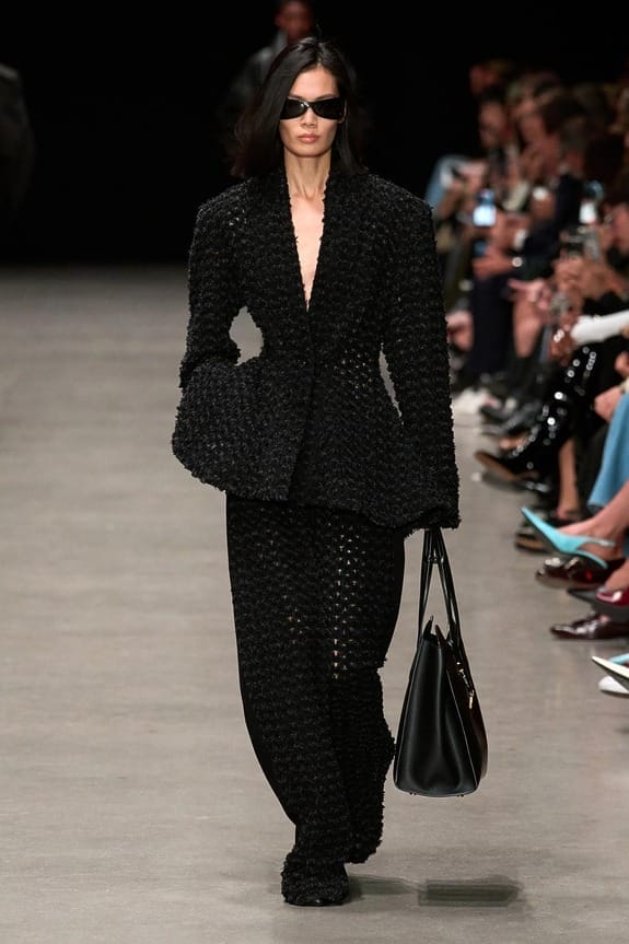 Model walking down runway in textured black outfit, wearing sunglasses and carrying a black bag, with audience in background.