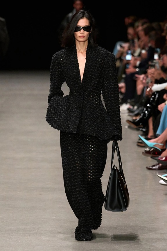 Model walking down runway in textured black outfit, wearing sunglasses and carrying a black bag, with audience in background.