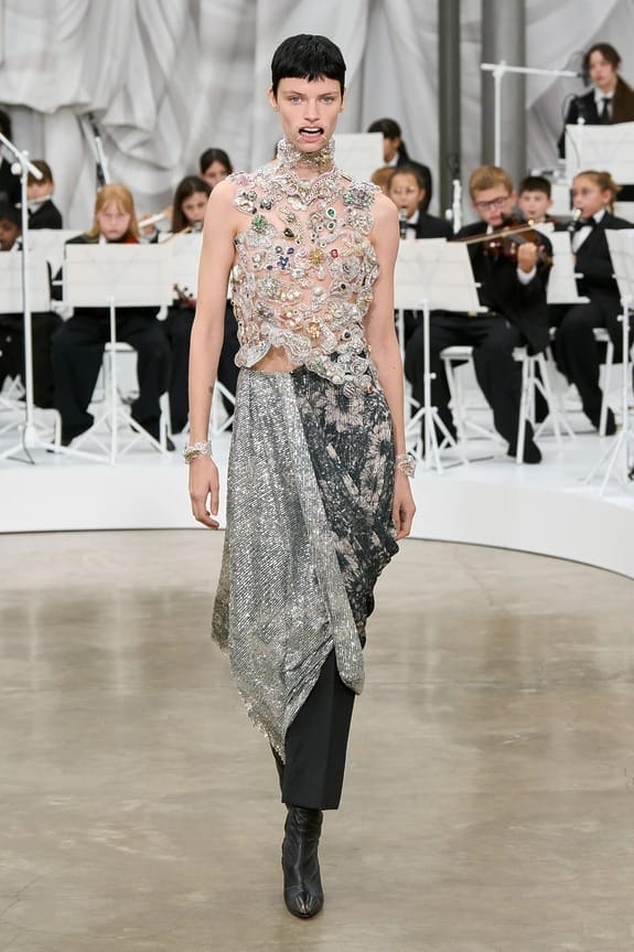 Model walking down runway in floral embellished top and silver skirt, children playing instruments in the background.