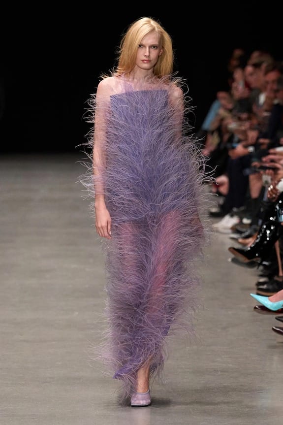Model walking runway in a textured purple feathered dress with audience in the background