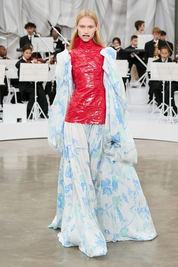 Model in a blue floral gown with red top walks a runway, orchestra playing in the background