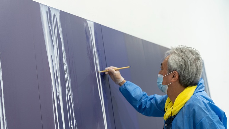 Artist painting vertical white streaks on a large purple canvas, wearing a blue jacket and mask.