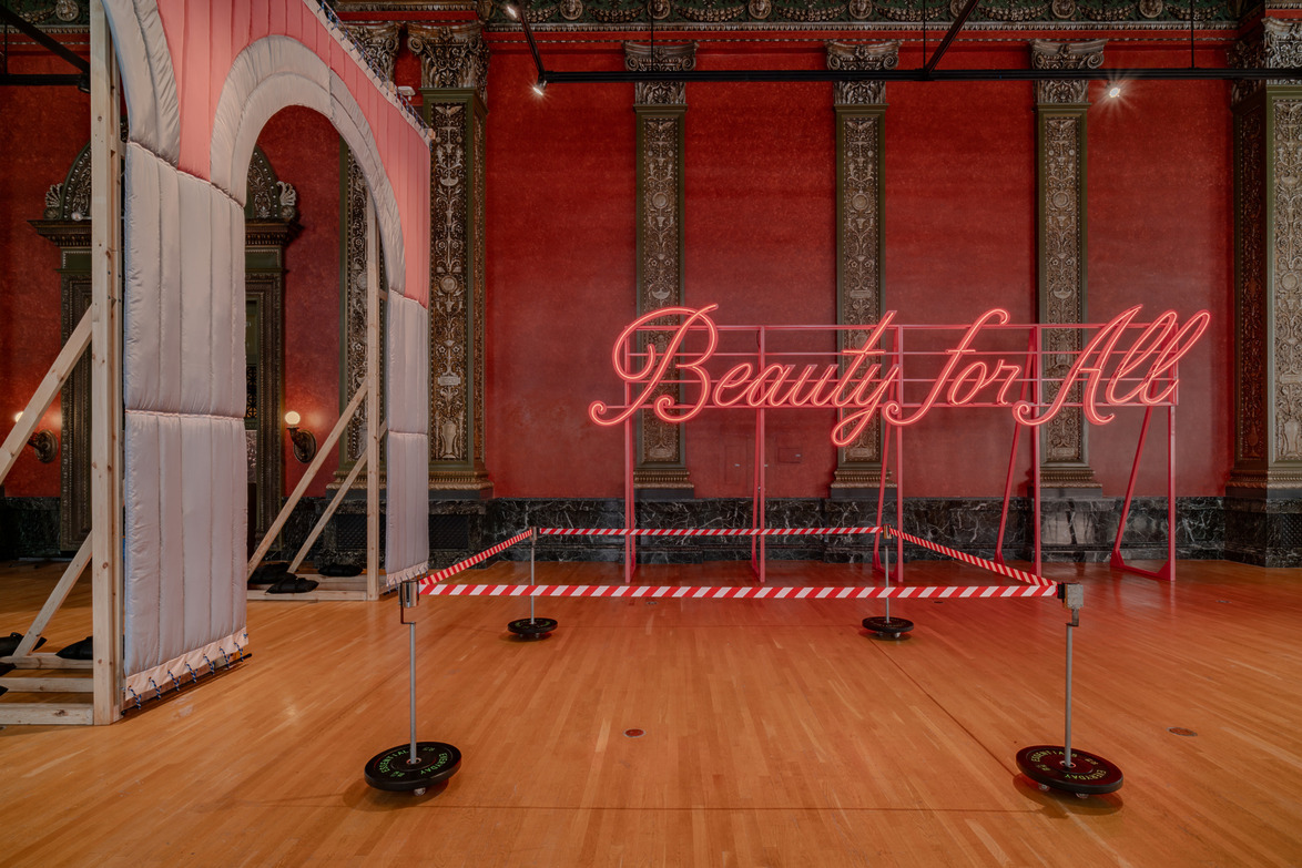 Neon sign reading "Beauty for All" in a red-walled room with ornate columns and a wooden floor.