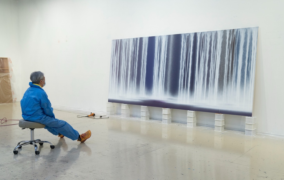 Artist sitting on a stool, observing large abstract painting of cascading waterfalls in a studio setting.