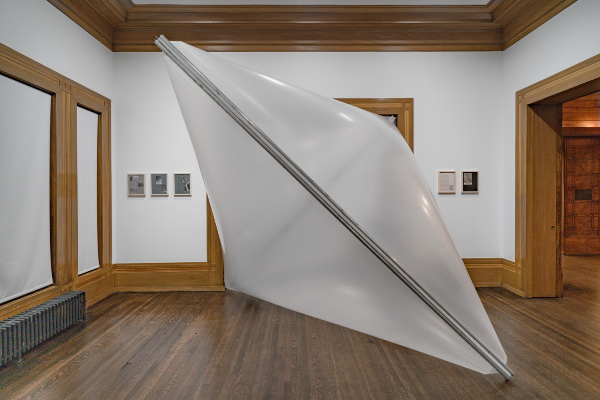 Large geometric sculpture resembling a folded sheet, displayed in a gallery room with wooden floors and framed pictures.
