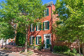 Historic three-story red brick house with green shutters and a tree-lined sidewalk on a sunny day.