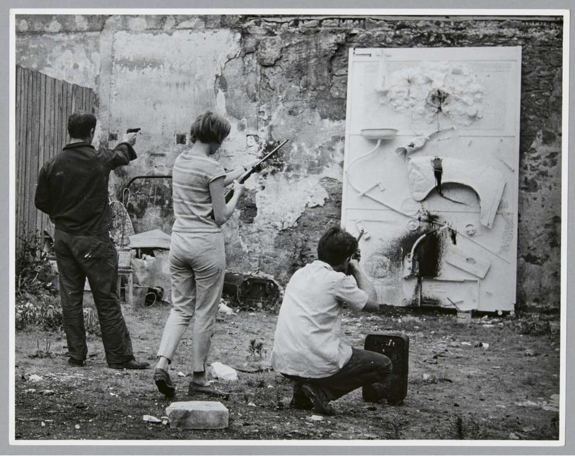 Three individuals engaging with a textured wall art piece in an outdoor setting, two painting and one photographing in grayscale.