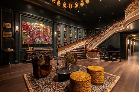 Elegant hotel lobby with grand staircase, colorful artwork, piano, velvet chairs, and ornate decor in a warmly lit setting.