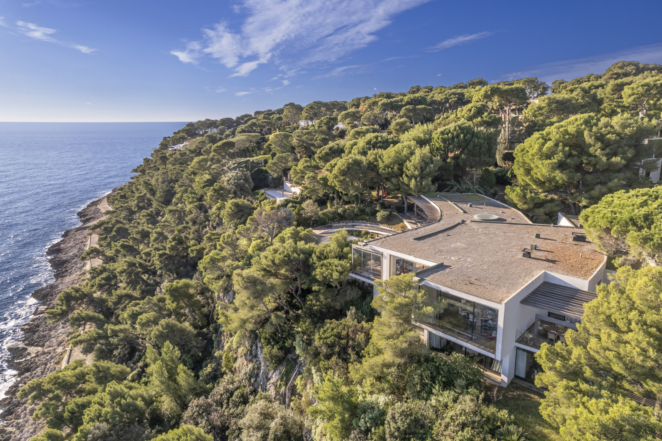 Modern house on a cliff surrounded by lush trees overlooking a vast ocean under a clear blue sky.