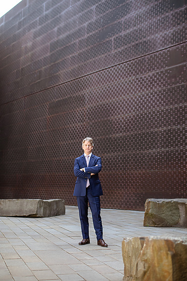 Man in blue suit standing with arms crossed in a modern architectural outdoor space.