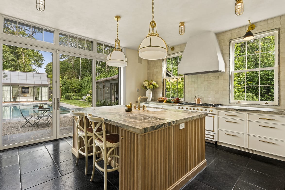 Modern kitchen with a marble island, large windows, and a view of a patio and pool. Bright and airy interior design.