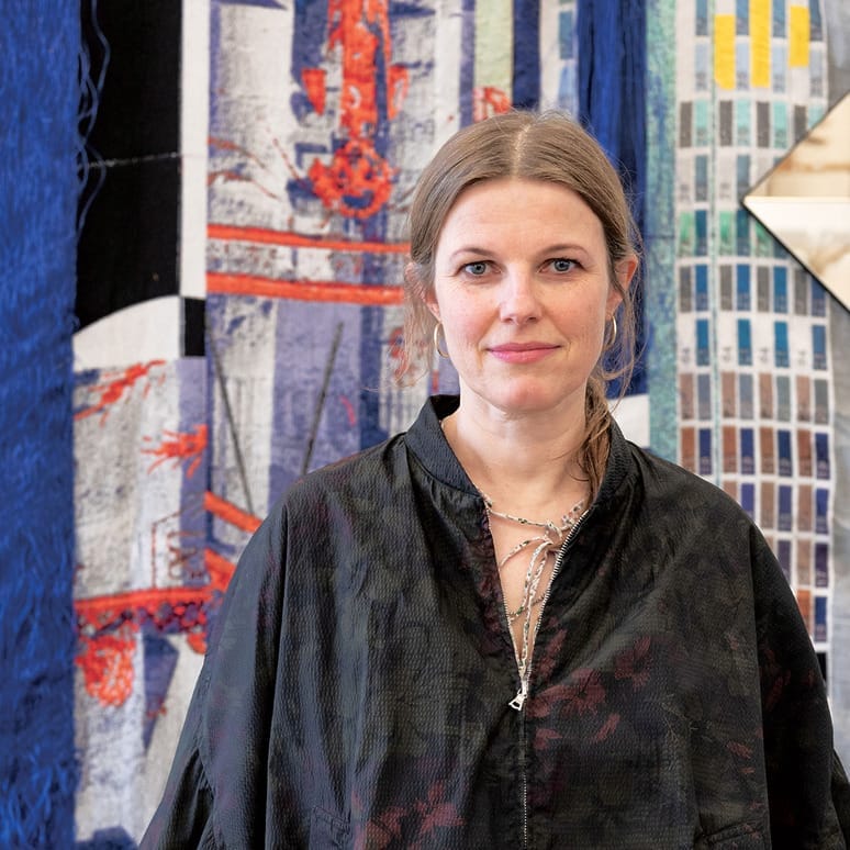 Person standing in front of a colorful, abstract artwork with various fabrics and textures in the background.