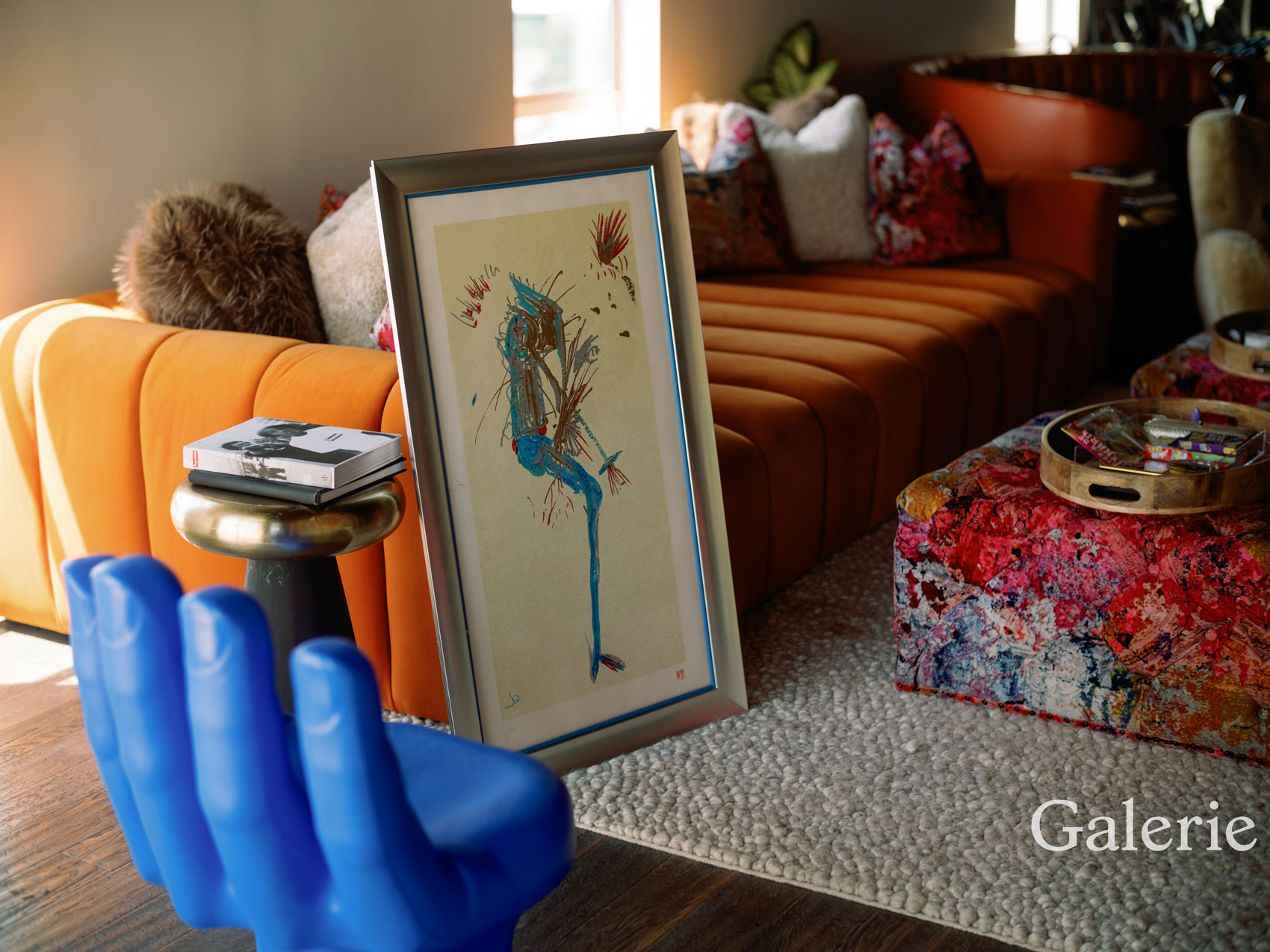 Framed artwork leaning against an orange sofa with plush cushions and a colorful coffee table in a cozy living room.