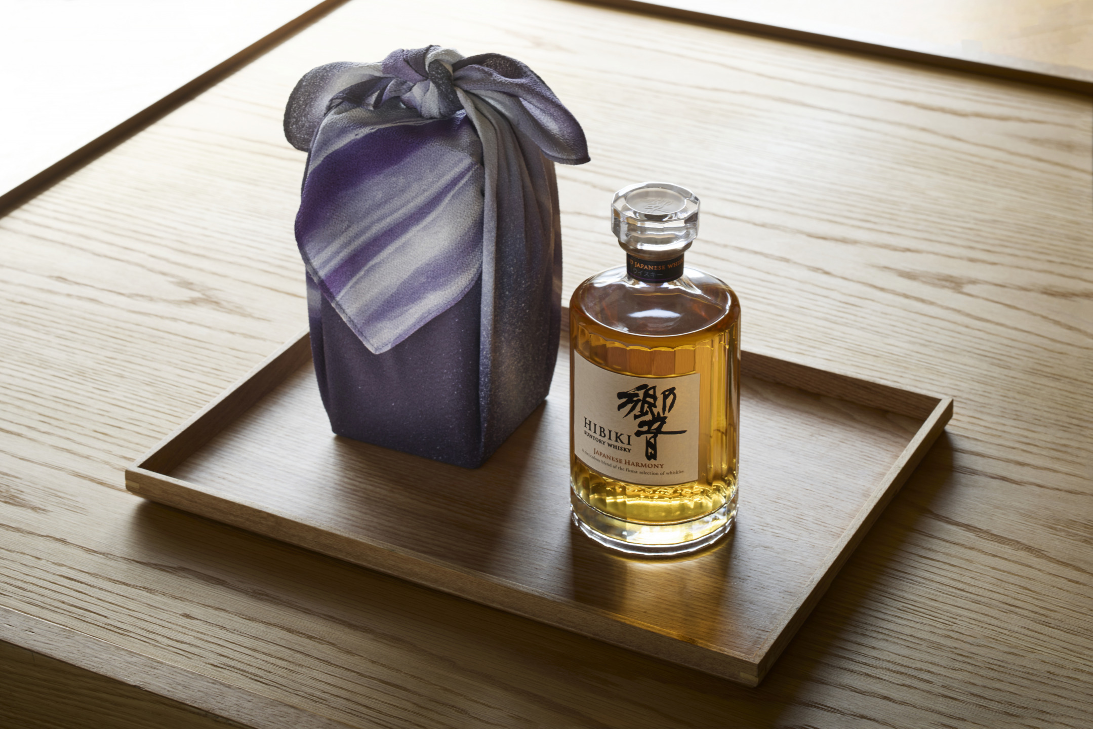 A bottle of whiskey with Japanese label on a wooden tray next to a purple wrapped gift on a wooden surface.