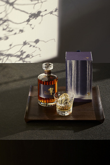 Bottle of amber liquid with a glass and box on a wooden tray, shadow of branches on wall in background.