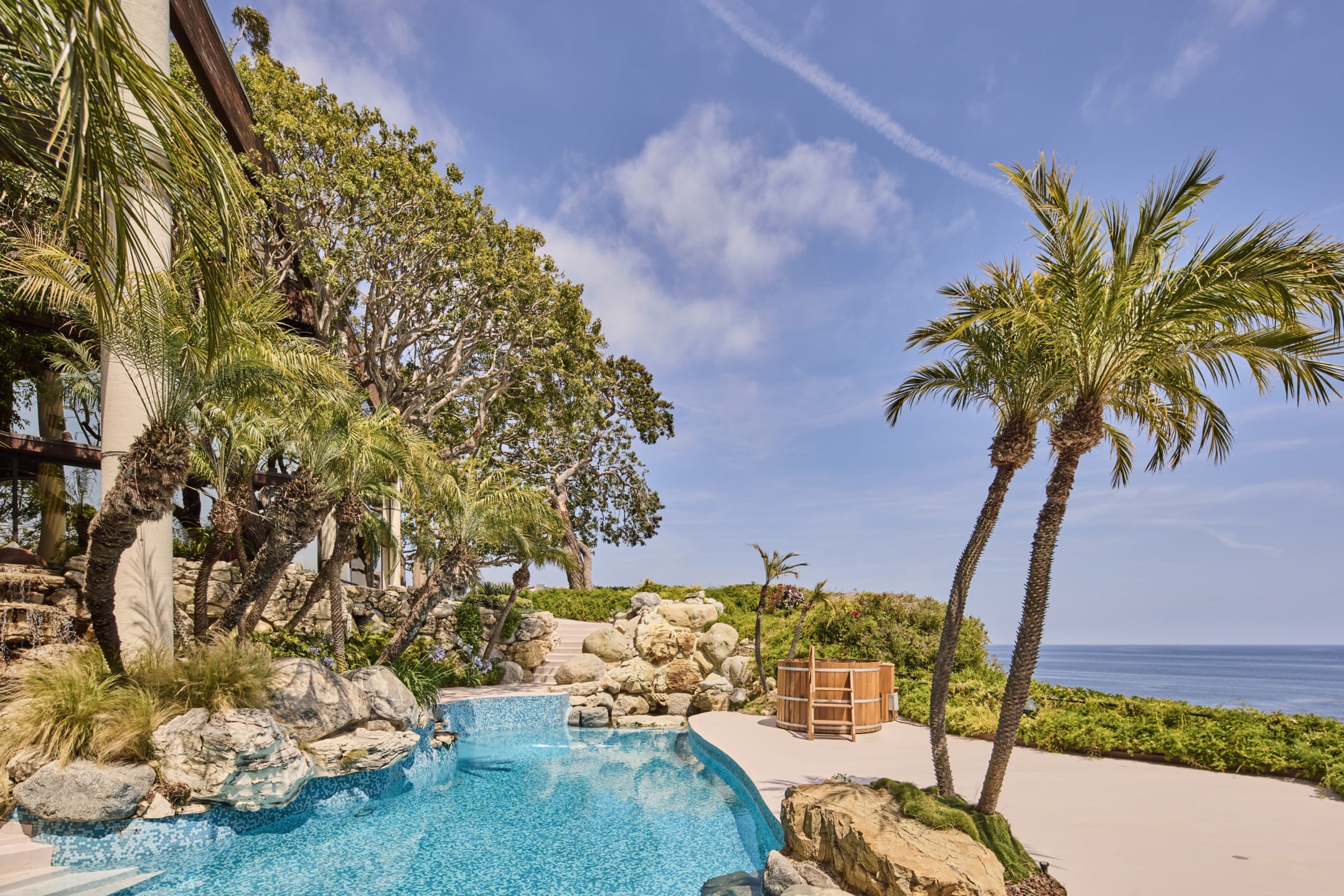 Tropical poolside with palm trees, ocean view, and clear blue sky. Rocky waterfall enhances the tranquil landscape.