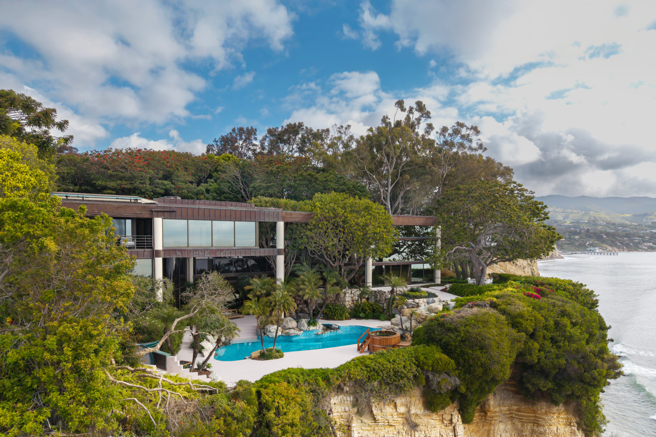 Luxury cliffside mansion with pool overlooking the ocean, surrounded by lush greenery and a bright blue sky with clouds