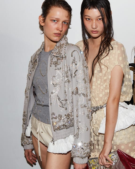 Two models in detailed sequined outfits, one wearing a silver jacket, the other in a beige dress, posing against a white background.