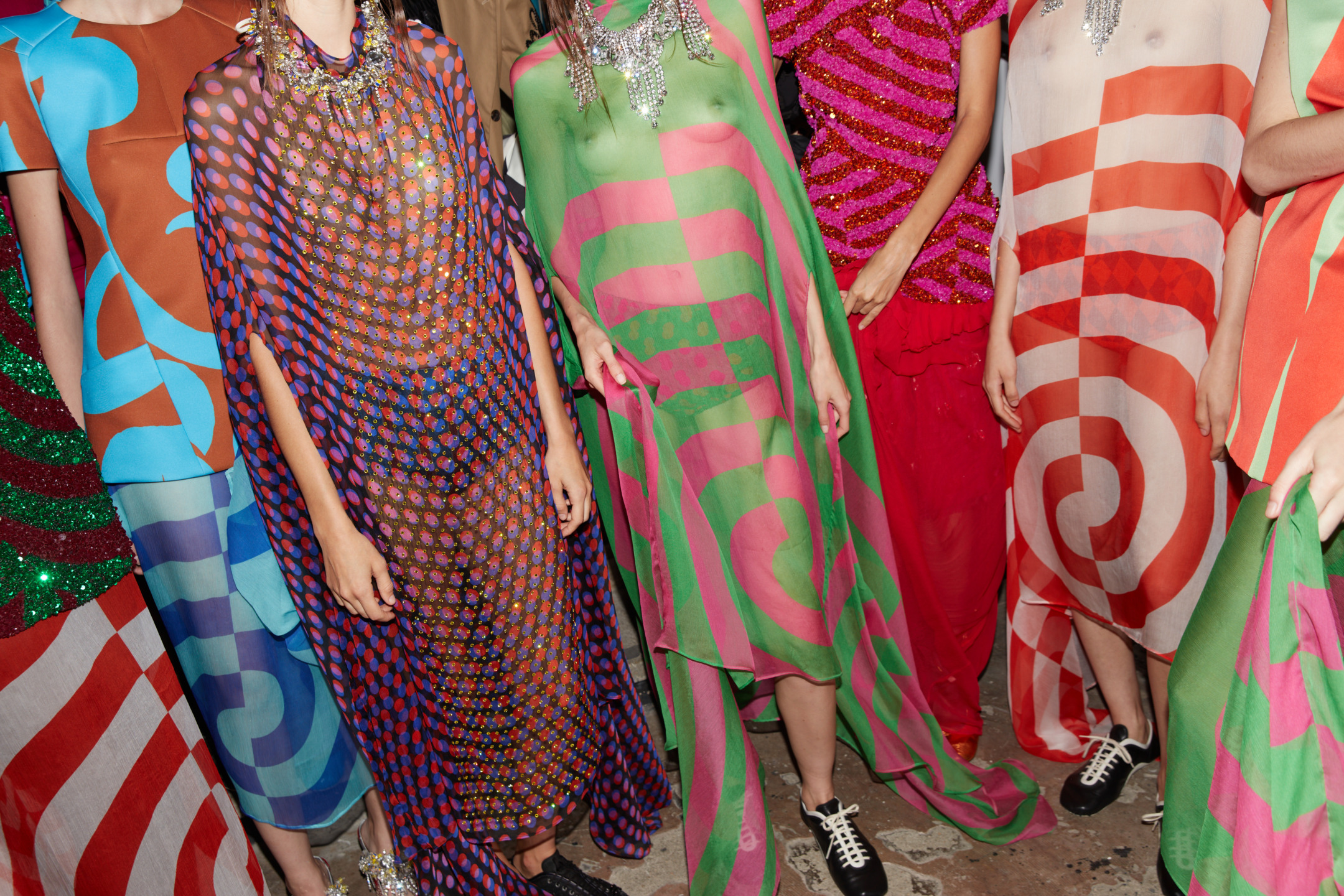 Models wearing colorful, swirling patterned dresses and sparkling accessories on a runway.