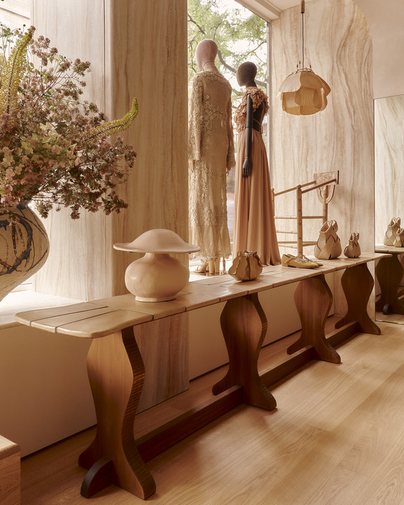 Fashion display with elegant dresses on mannequins, styled with a wooden bench and floral arrangement in a warmly lit boutique.
