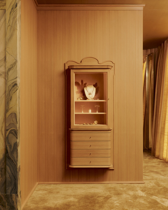 Elegant wooden jewelry cabinet with glass door displaying necklaces and rings against a marble and wood-paneled backdrop.