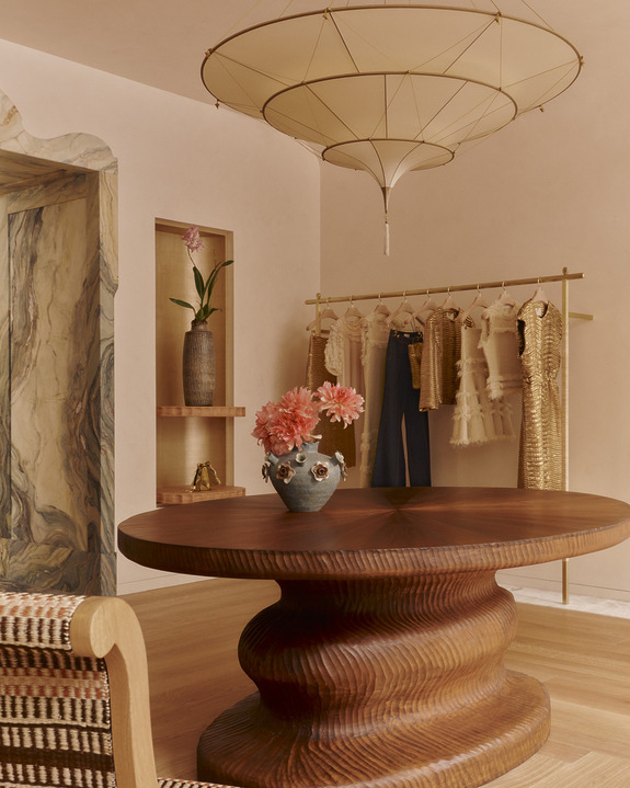 Elegant interior with round wooden table, floral vase, hanging clothes, and unique overhead light fixture.