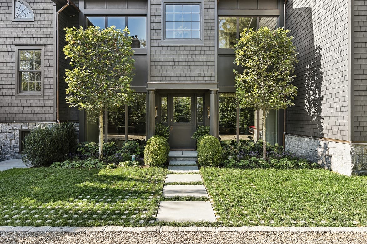 Modern house facade with a pathway, green lawn, and manicured bushes, framed by two tall trees, under a clear sky.