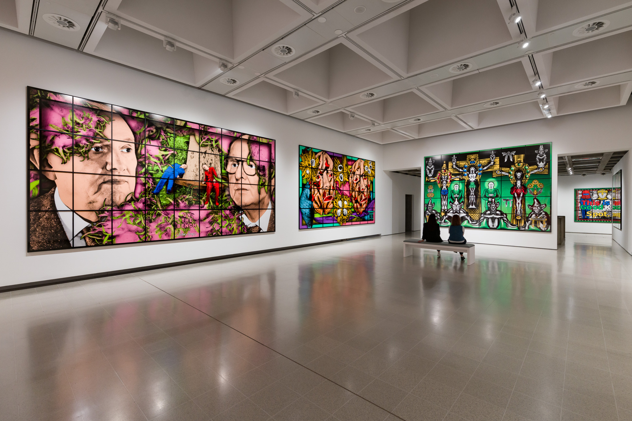 Art gallery interior featuring large colorful modern artworks on walls, with two visitors seated on a bench observing.