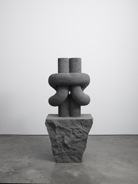 Abstract stone sculpture on a textured base in a minimalist gallery setting.