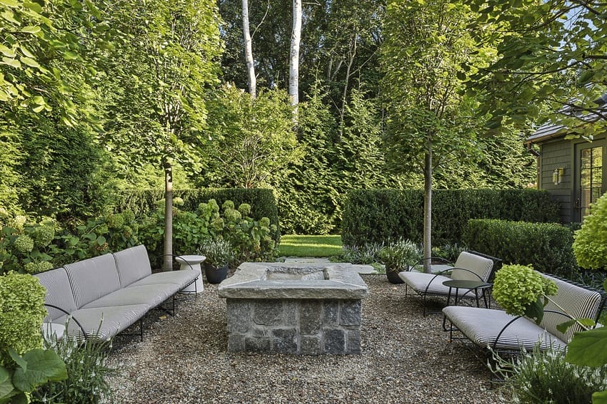 Outdoor garden area with stone fire pit, surrounded by trees, hedges, and cushioned benches on a gravel patio.