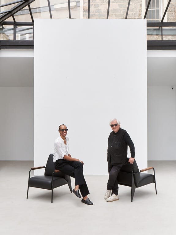 Two people sitting on modern black chairs in a minimalistic room with large windows and a white backdrop.