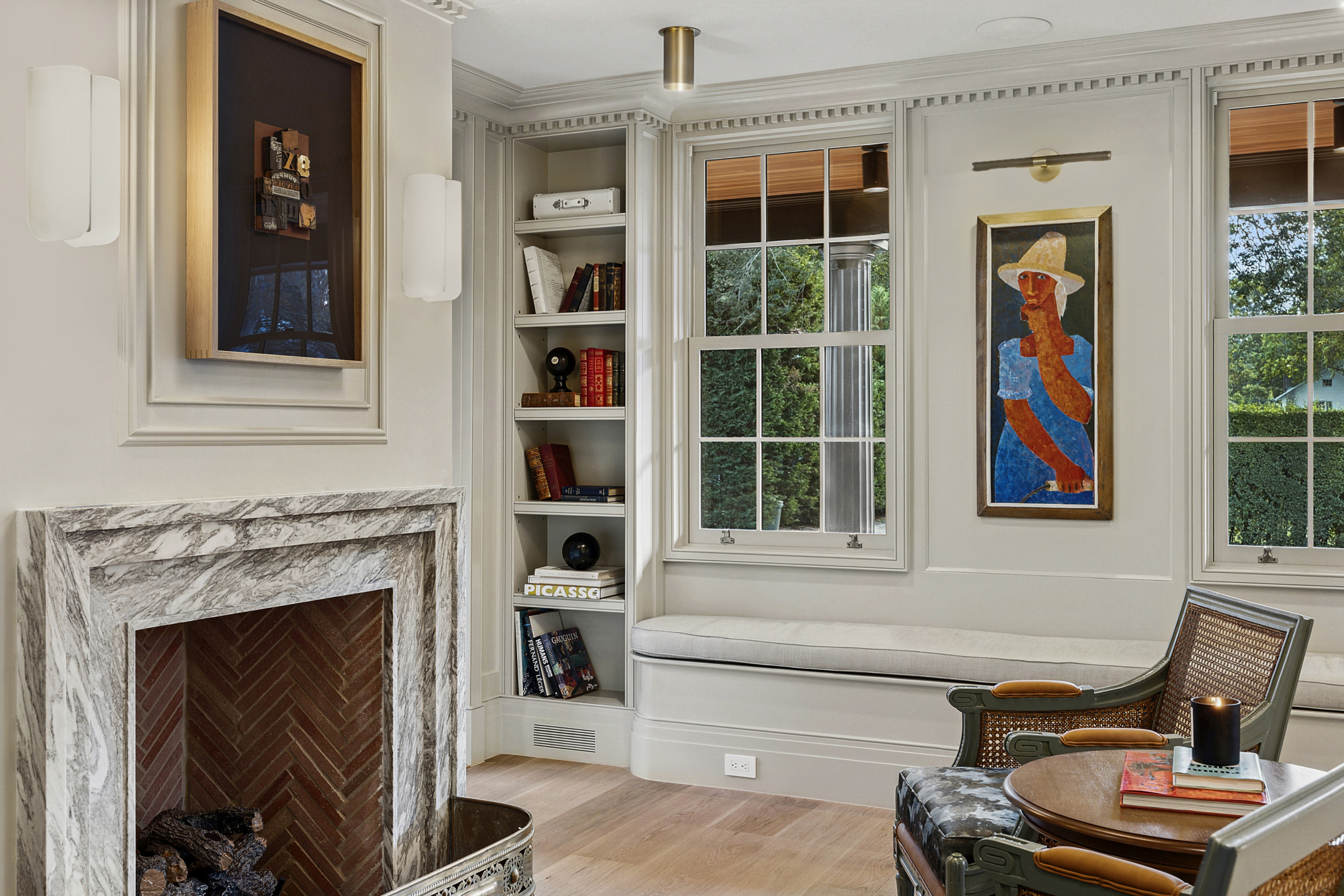 Cozy living room with marble fireplace, artwork on walls, bookshelves, and large windows with garden view.
