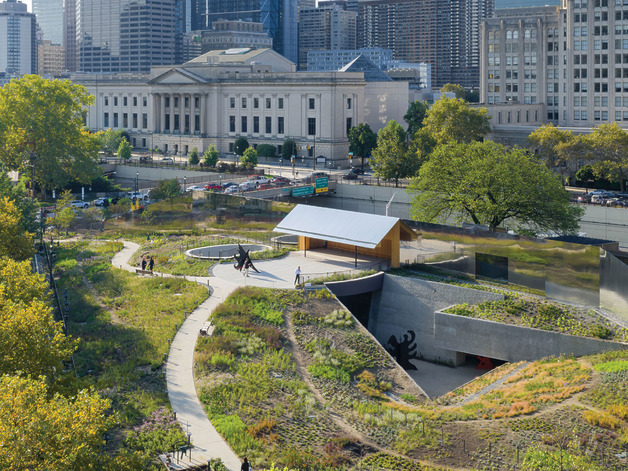 Urban park with winding paths, greenery, art installations, and city buildings in the background on a sunny day.