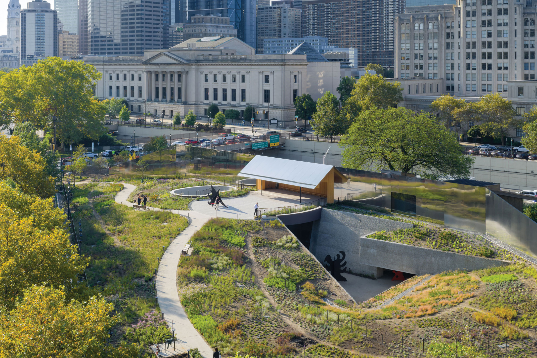 Urban park with winding paths, greenery, art installations, and city buildings in the background on a sunny day.