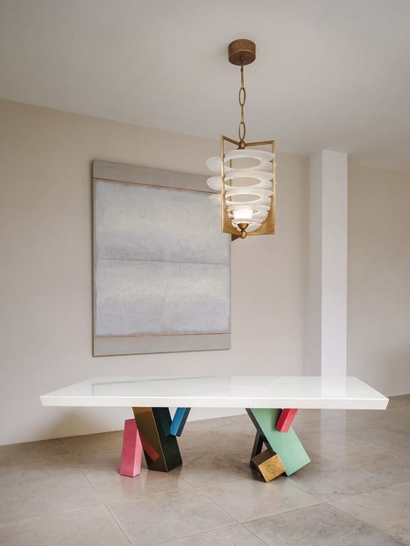 Modern dining room with a colorful abstract table, geometric chandelier, and a large minimalist painting on a beige wall.