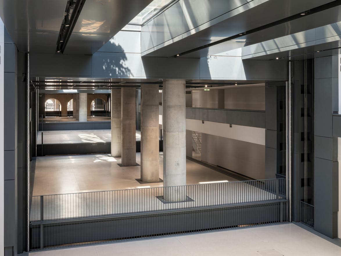 Interior view of a modern building with concrete pillars, large windows, and a sleek design under bright natural lighting.