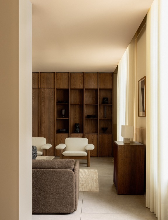 Minimalist living room with a neutral color palette, featuring wooden shelves, white chairs, a brown sofa, and soft lighting.