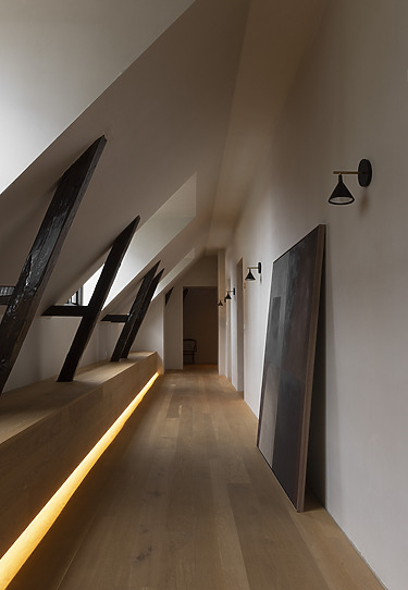 Minimalist attic hallway with wooden floor, modern wall lights, exposed beams, and leaning artwork.