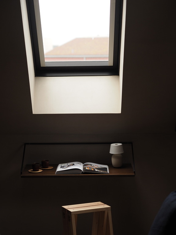 Minimalist workspace under a skylight with an open magazine, two mugs, a lamp, and a wooden stool.