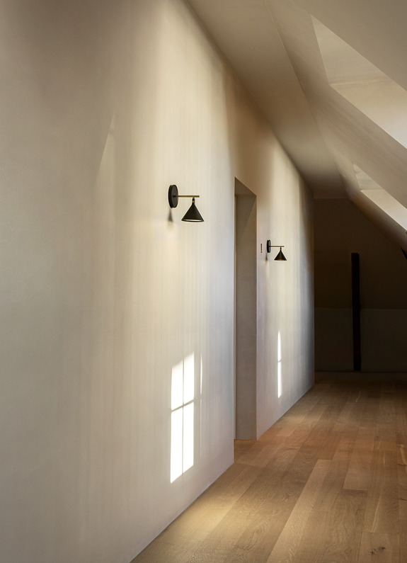 Minimalist hallway with warm wooden floor and modern black wall sconces, illuminated by sunlight streaming through a window.