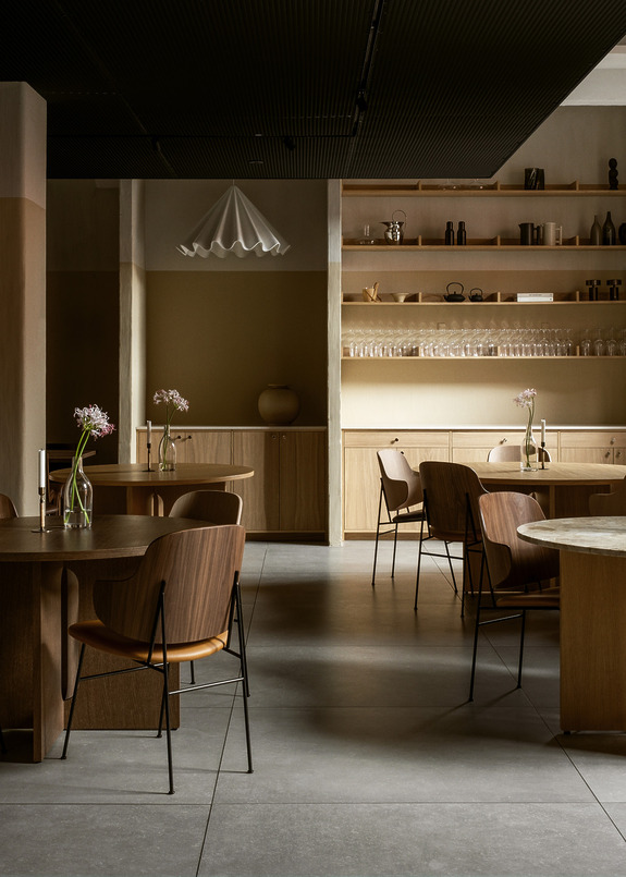 Modern restaurant interior with wooden tables, chairs, and minimalist decor. Shelving with glasses and ceramics in the background.