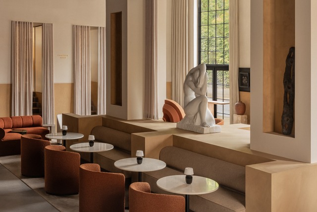 Modern indoor lounge with warm tones, circular tables, orange chairs, and a large artistic sculpture by large windows.