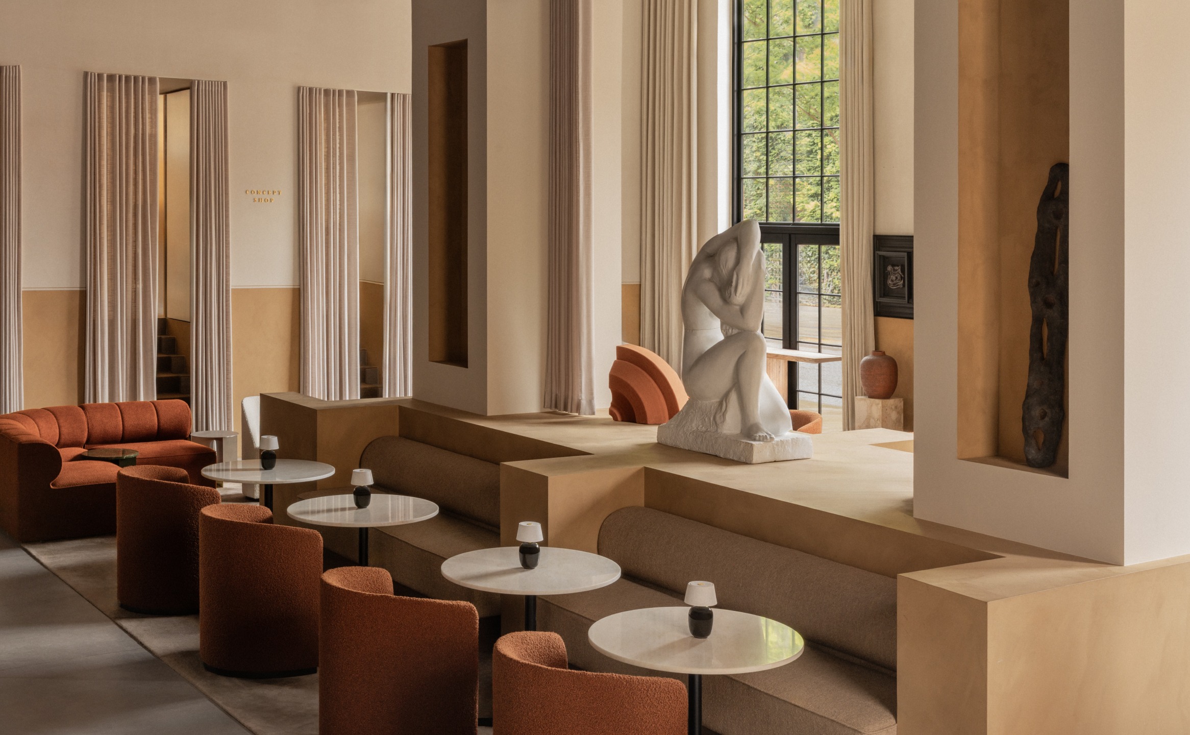 Modern indoor lounge with warm tones, circular tables, orange chairs, and a large artistic sculpture by large windows.