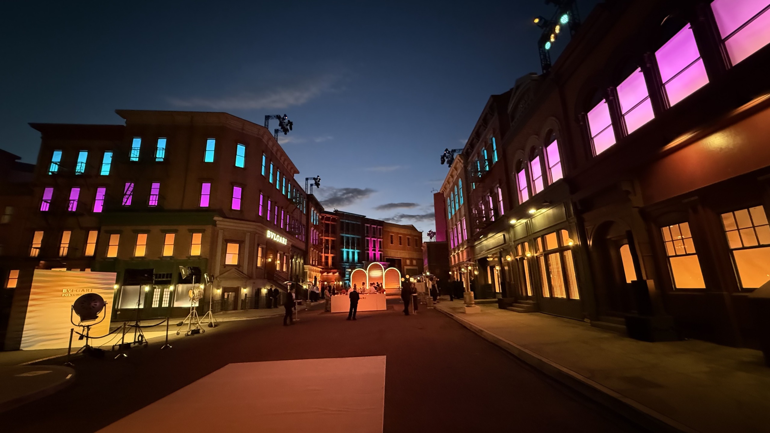 Street scene at dusk with buildings illuminated by colorful lights, people walking, and a dramatic twilight sky.