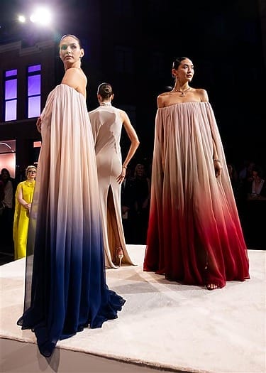Models showcase elegant ombre gowns on a runway with blue and red hues, with an audience watching in the background.