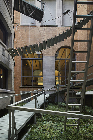 Modern architectural installation with suspended metal walkways and large arched windows in a brick and concrete courtyard.