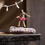 A small handmade doll in a pink skirt balances on a striped cushion with a vintage backdrop.