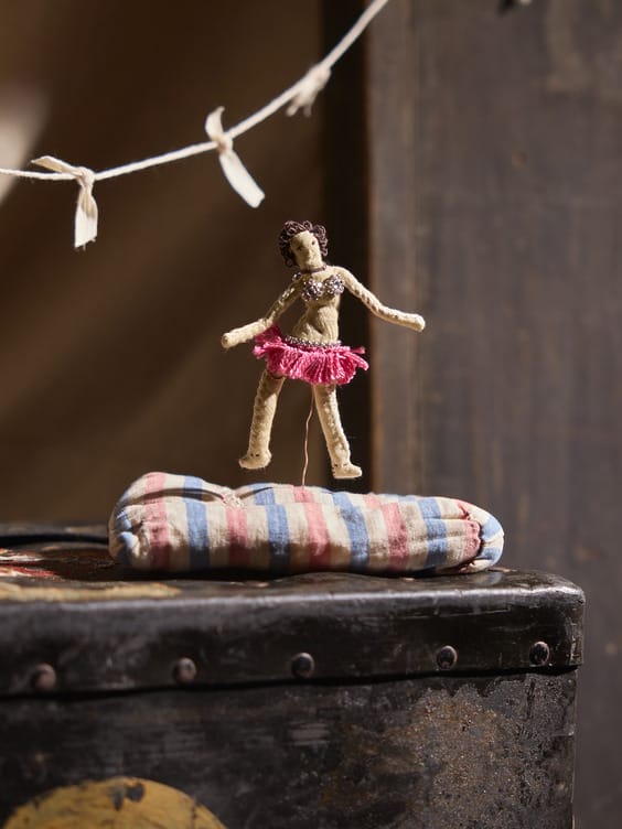A small handmade doll in a pink skirt balances on a striped cushion with a vintage backdrop.
